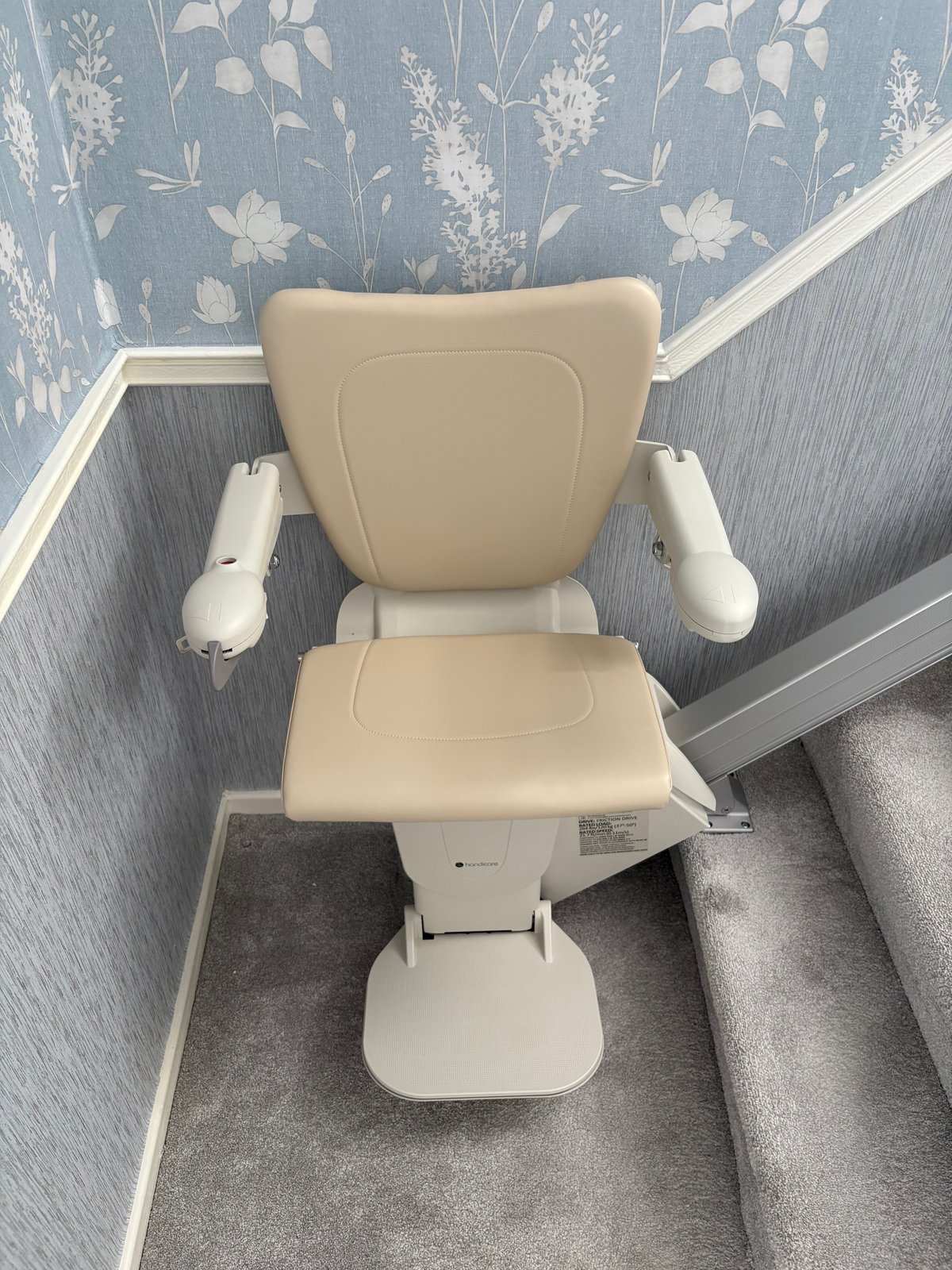 New straight stairlift with a beige seat, armrests and footrest, fitted on a straight rail beside carpeted stairs